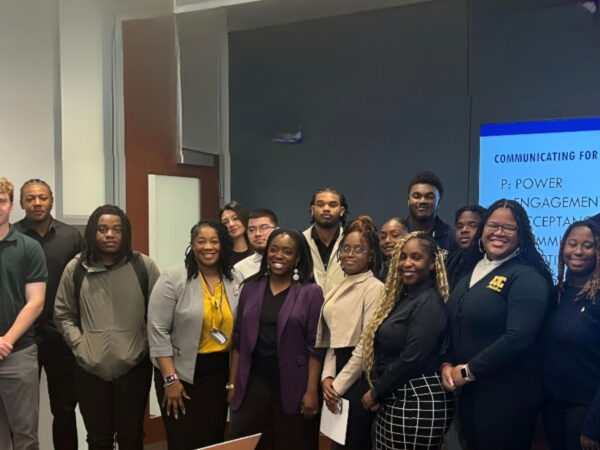 Group of diverse young professionals and students posing for a photo in front of a blue presentation screen in a conference space.