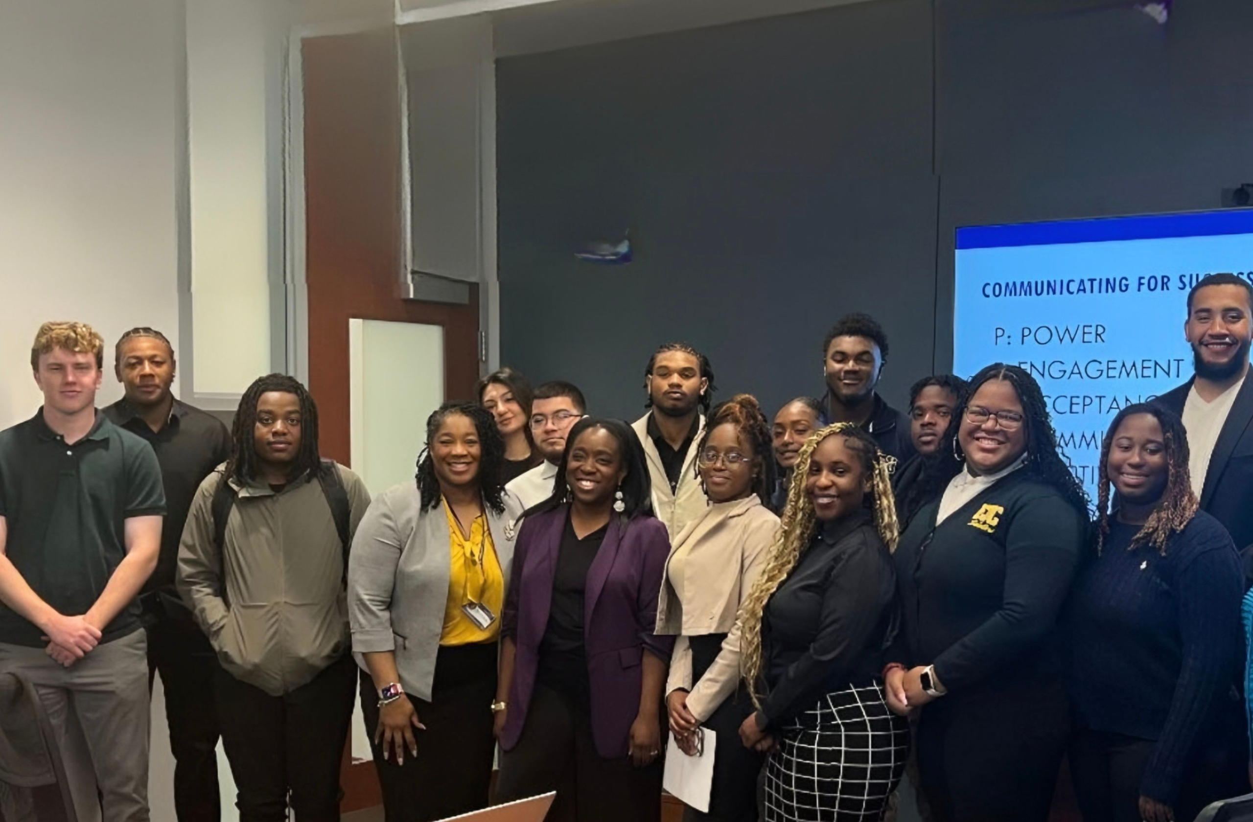 Group of diverse young professionals and students posing for a photo in front of a blue presentation screen in a conference space.