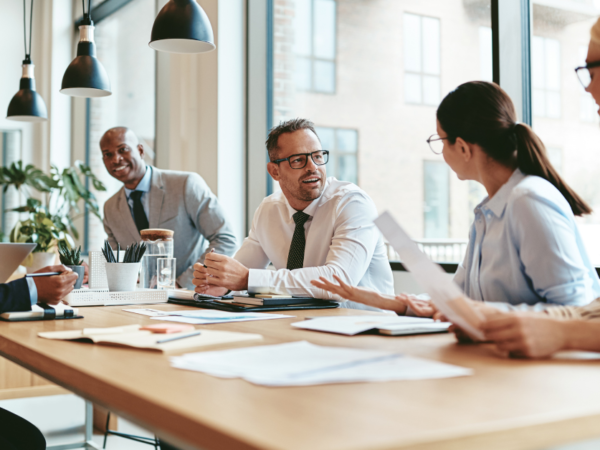 Diverse group of coworkers in a modern office discussing documents around a wooden conference table.