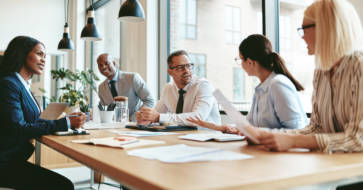 Diverse group of coworkers in a modern office discussing documents around a wooden conference table.